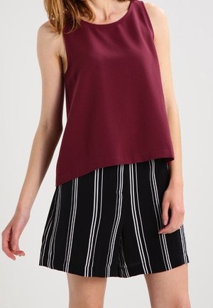 Femme portant un haut bordeaux sans manches et un short noir à rayures blanches verticales, debout devant un fond uni.