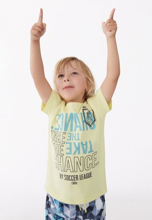 Camiseta de manga corta amarilla clara con texto azul que dice "TOMA EL RIESGO" y un emblema de fútbol. Combinada con pantalones cortos de baño con estampado.