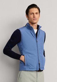 Muž v světle modré bezrukávé vestě na zip přes tmavé tričko s dlouhým rukávem a světlé kalhoty, stojící před jednobarevným pozadím.