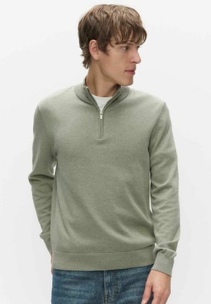 Młody mężczyzna w jasnooliwkowym swetrze z zamkiem przy szyi i niebieskich dżinsach, patrzący w prawo na jednolitym białym tle.