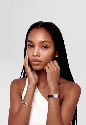 Mujer con largas trenzas que lleva una blusa blanca, pendientes y collar de perlas, un reloj con correa negra y dos pulseras, tocándose suavemente el rostro.