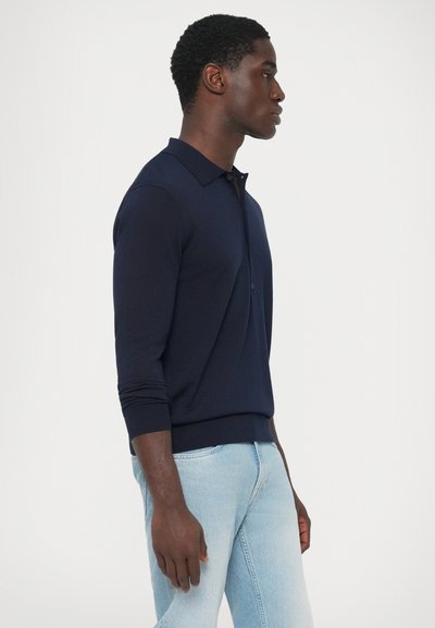 Giovane in polo a maniche lunghe blu navy e jeans azzurri che sta in piedi di lato contro uno sfondo bianco semplice.