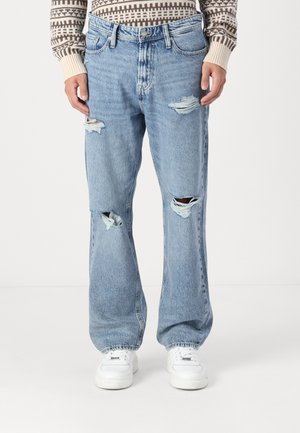 Vaqueros desgastados de color azul claro con rotos prominentes en las rodillas, un ajuste holgado y un dobladillo deshilachado, combinados con zapatillas blancas sobre un fondo neutro.
