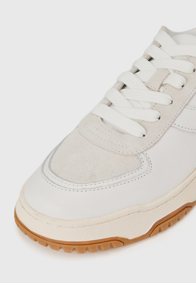 Weiße Leder Sneaker mit Wildlederakzenten. Merkmale sind flache weiße Schnürsenkel, gummierte Sohle und gepolsterte Zunge. Abgerundete Zehenform.