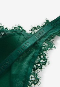 Zelena čipkana grudnjak s složenim čipkastim rubovima i satenskim pojasom s oznakom "Lipsy." Materijal se čini mekim i teksturiranim, naglašavajući delikatnu izradu.
