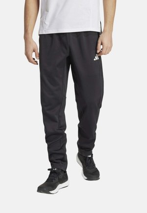 Persona con pantalones deportivos negros con un logotipo blanco en el muslo, zapatillas negras y una camiseta blanca sobre un fondo liso.