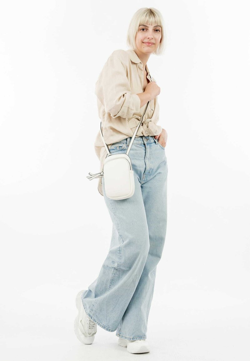 Camisa beige de manga larga con botones, jeans de pierna ancha azul claro y zapatillas blancas. Un pequeño bolso blanco cruzado con un detalle de cremallera.