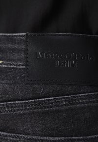 Crni traper tkanina sa glatkom teksturom, s pravokutnom kožnom oznakom utisnutom s natpisom "Marc O'Polo DENIM." Istaknuti šavovi su vidljivi.