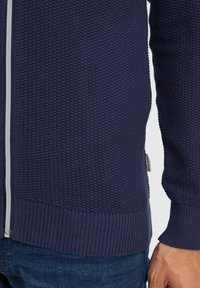 Námornícky texturovaný sveter na zips s rebrovaným lemom a manžetami, so zvýšeným vzorom a malou značkovou štítkom na boku.