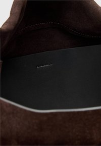 Binnenkant van bruin suède met een gladde zwarte leren basis, voorzien van een subtiel geperst logo. Afgeronde hoeken en een strak, eenvoudig ontwerp.