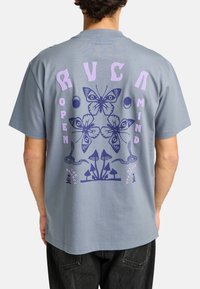 Camiseta de algodón gris con un gran gráfico en la parte posterior que presenta diseños de mariposas moradas y azules, champiñones y el texto "MENTE ABIERTA" con elementos decorativos.