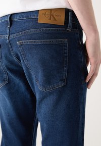 Tamnoplave traperice klasičnog kroja, s smešnom kožnom etiketom na zadnjem delu struka na kojoj je ugravirano "Calvin Klein Jeans."