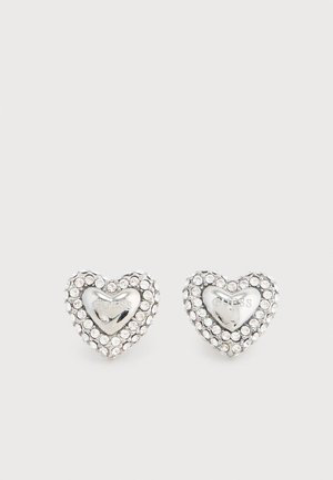 Silver heart-shaped stud earrings with clear rhinestones surrounding a polished center engraved with "GUESS" brand name.
