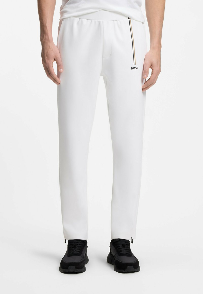 Homme portant un jogging blanc BOSS avec une bande verticale et des baskets noires, debout sur un fond blanc.