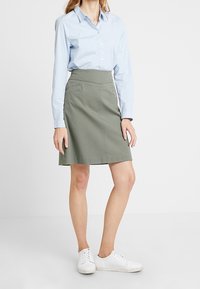 Gonna A-line khaki con vita alta, tasche laterali e cuciture a contrasto. Abbinata a una camicia botton-down azzurra chiara e sneakers bianche.