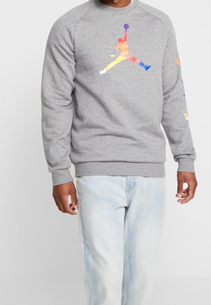 Osoba nosząca szarą bluzę z wielokolorowym logo Jumpman i jasnoniebieskie dżinsy, stojąca na jednolitym jasnym tle.