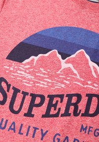 Camiseta de color rojo heather con un gráfico de montañas, olas del océano y la palabra impresa "SUPERD" en texto negrita azul marino.