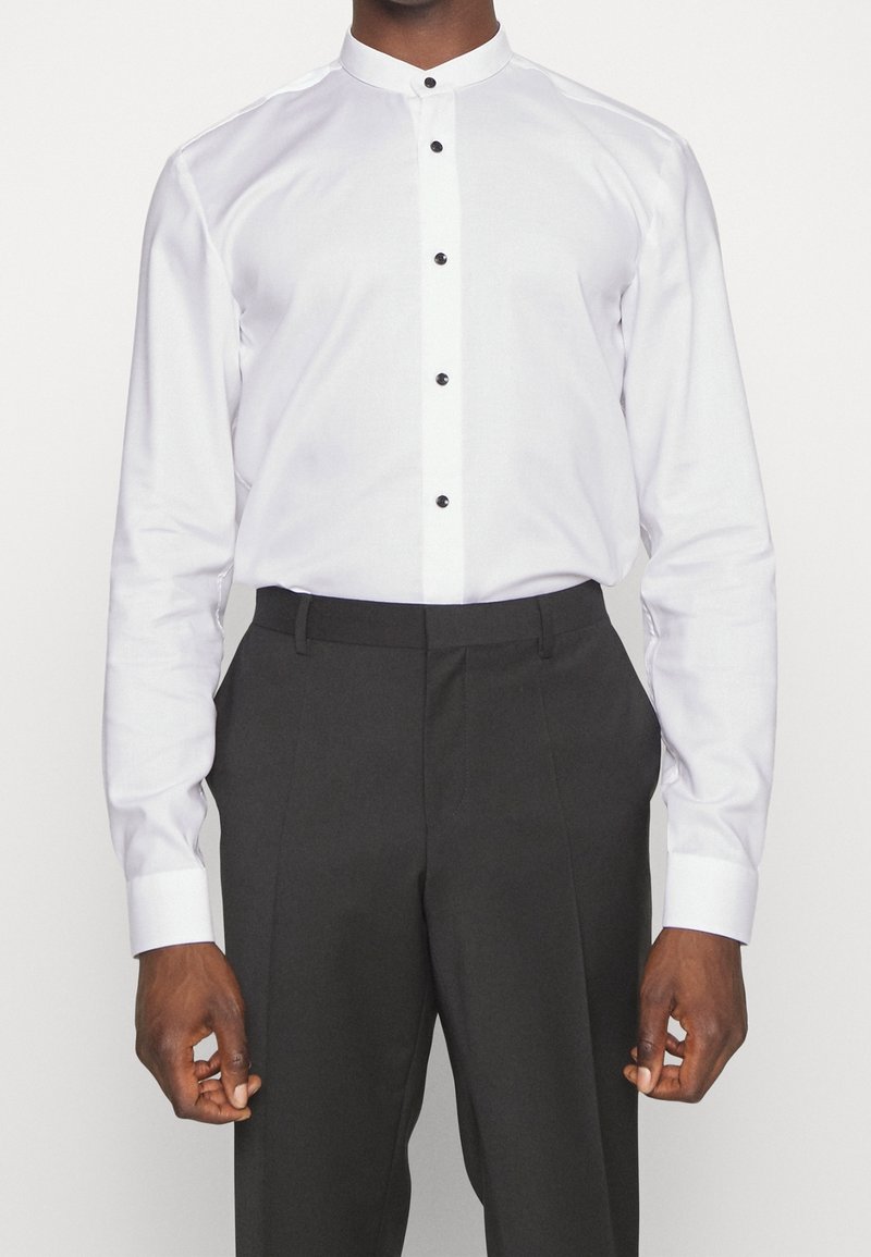 Uomo che indossa una camicia bianca con colletto alla mandarino e bottoni neri, insieme a pantaloni neri su misura, su uno sfondo neutro.