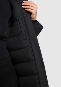Sorte pufferjakke med en quiltet design, der viser et glat skalmateriale, sidelommer og knaplukning.