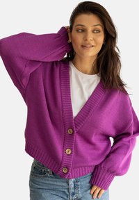 Magenta, oversize dziergany kardigan z dekoltem w serek, z drewnianymi guzikami i ściągaczami przy mankietach. Połączony z białą koszulką i szortami dżinsowymi.