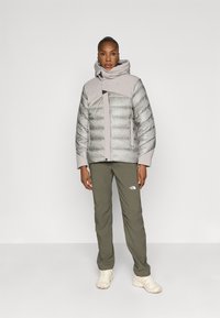 Giacca grigia chiara e imbottita con cappuccio, caratterizzata da un motivo a rombi e chiusura a zip. Abbinata a pantaloni verde oliva e scarpe chiare.