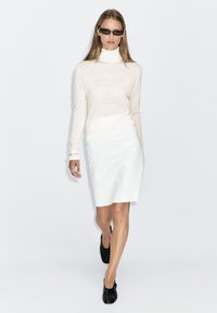 Lichtbeige coltrui, witte kokerrok en zwarte instapschoenen; model draagt ook een donkere zonnebril; minimalistisch ontwerp.