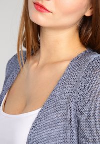 Světlý šedý pletený cardigan s volným otevřeným střihem, texturovanou látkou a širokými rukávy, nošený přes bílý top.