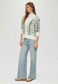 Weißer V-Ausschnitt-Cardigan mit grünem und schwarzem Karomuster, kombiniert mit hellblauen Weitbeinen Jeans und braunen Plateauschuhen.