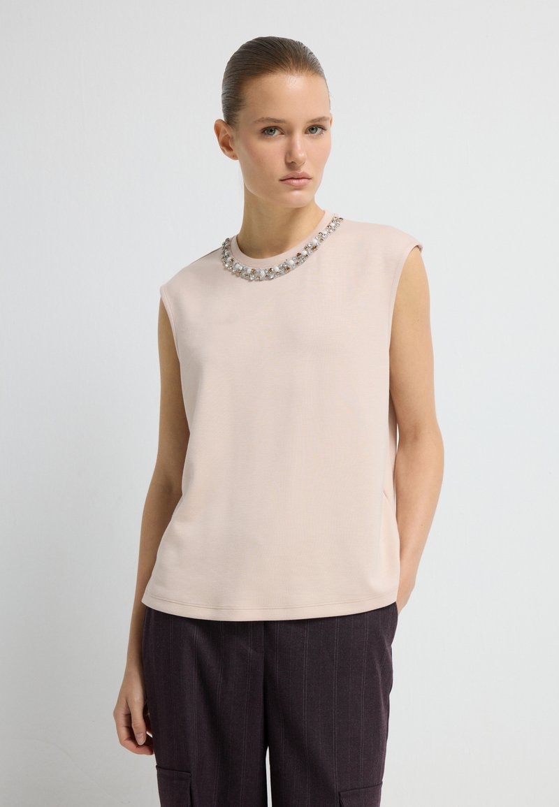 Junge Frau mit zurückgebundenem Haar trägt ein ärmelloses beiges Oberteil mit einem Halsband aus Perlen und Perlen, kombiniert mit dunkel gestreiften Hosen und steht aufrecht.