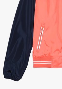 Bomber bunda s blokováním barev v korálové a námořnické modři; obsahuje zipovou kapsu, elastické manžety a pruhované žebrování na lemu. Hladká textilie.