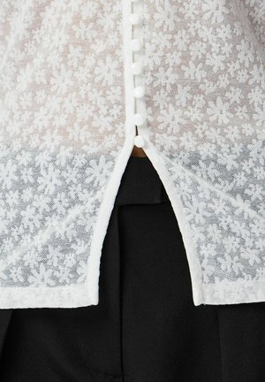 Blusa blanca translúcida de encaje floral con pequeños botones redondos en el centro sobre tela negra visible a través de una abertura en la parte inferior.