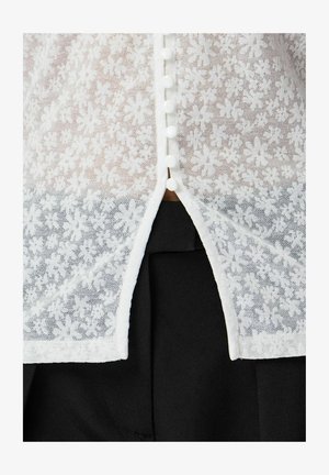 Bluza albă de dantelă florală semi-transparentă cu nasturi mici rotunzi pe centrul feței, peste material negru vizibil printr-o crăpătură jos.