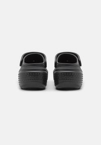 Paire de sabots noirs à enfiler avec des brides au talon, surface texturée visible et logo à l'arrière, vus de dos sur fond blanc.