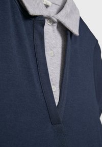 Pull en col V bleu marine superposé sur une chemise boutonnée gris clair avec un col et des boutons visibles.