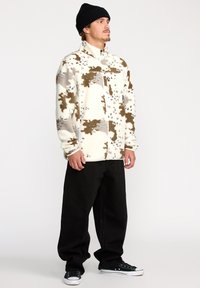 Veste en polaire blanche avec des taches brunes et grises, dotée d'un col haut et d'une fermeture éclair à l'avant. Associée à un pantalon large noir et des baskets.