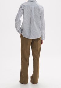 Gestreiftes Hemd in Weiß und Marine mit einem Rückenteil-Detail, kombiniert mit hochgeschnittenen braunen Hosen. Glatte Textur, lange Ärmel, Knöpfe an den Manschetten.