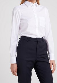 Camicia bianca a maniche lunghe con colletto classico e una singola tasca sul petto, abbinata a pantaloni su misura di un blu navy scuro.
