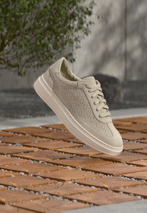 Zapatilla de ante beige con patrón geométrico perforado, cordones beige y suela gruesa, flotando sobre un suelo de madera y piedra al aire libre.