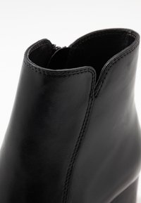 Černá kožená kotníková bota s elegantním tvarem, se šitým švem podél strany a zipovým uzávěrem na zadní straně. Hladký povrch.
