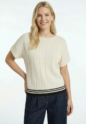 Femme souriante aux cheveux blonds portant un pull en maille beige à manches courtes avec des rayures noires et blanches à l'ourlet et un pantalon bleu foncé.