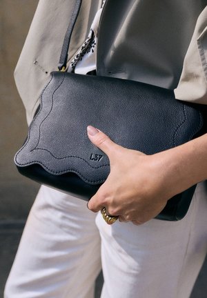 Hand hält eine schwarze, strukturierte Leder-Umhängetasche mit gewellten Kanten, trägt einen beigen Blazer und weiße Hose, goldener Ring am Finger.