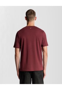 Tumši sarkana īsroku T-krekls no mīksta auduma ar apaļu kaklu un smalku logo virs kakla, kombinēts ar tumšām džinsu biksēm.