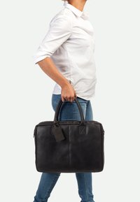 Schwarze Lederakte mit glatter Textur, doppelten oberen Griffen, einem Etikett und einem Reißverschluss, gehalten von einer Person in Jeans.