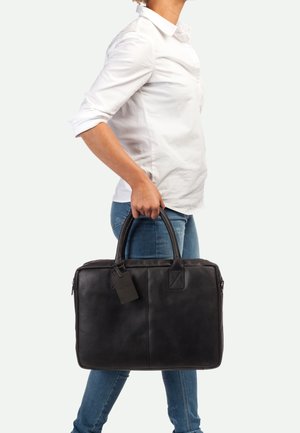 Une mallette en cuir noir avec une texture lisse, deux poignées supérieures, une étiquette et une fermeture éclair, tenue par une personne en jeans.