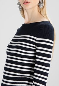 Maglione a righe navy e bianche con scollatura a barca. Maniche lunghe e design aderente, abbinato a grandi orecchini decorativi.