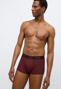 Cuecas boxer vermelhas com cintura elástica preta com o logo "HUGO", padrão geométrico em vermelho e preto, design ajustado ao corpo, textura suave.
