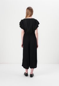 Black jumpsuit with short, ruffled sleeves, textured bodice, elastic waist, and wide leg design, worn with black flat shoes.