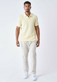 Camisa polo de tejido acanalado en amarillo claro con cuello, combinada con pantalones beige y zapatillas blancas. El modelo está de pie frente a un fondo sencillo.