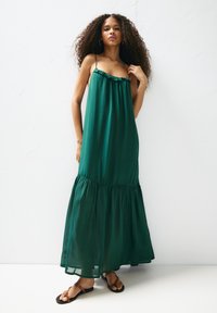 Rochie maxi verde, fluidă, cu bretele subțiri, decolteu volanat și fustă etajată. Stilizată cu sandale plate, așezată pe un fundal alb simplu.