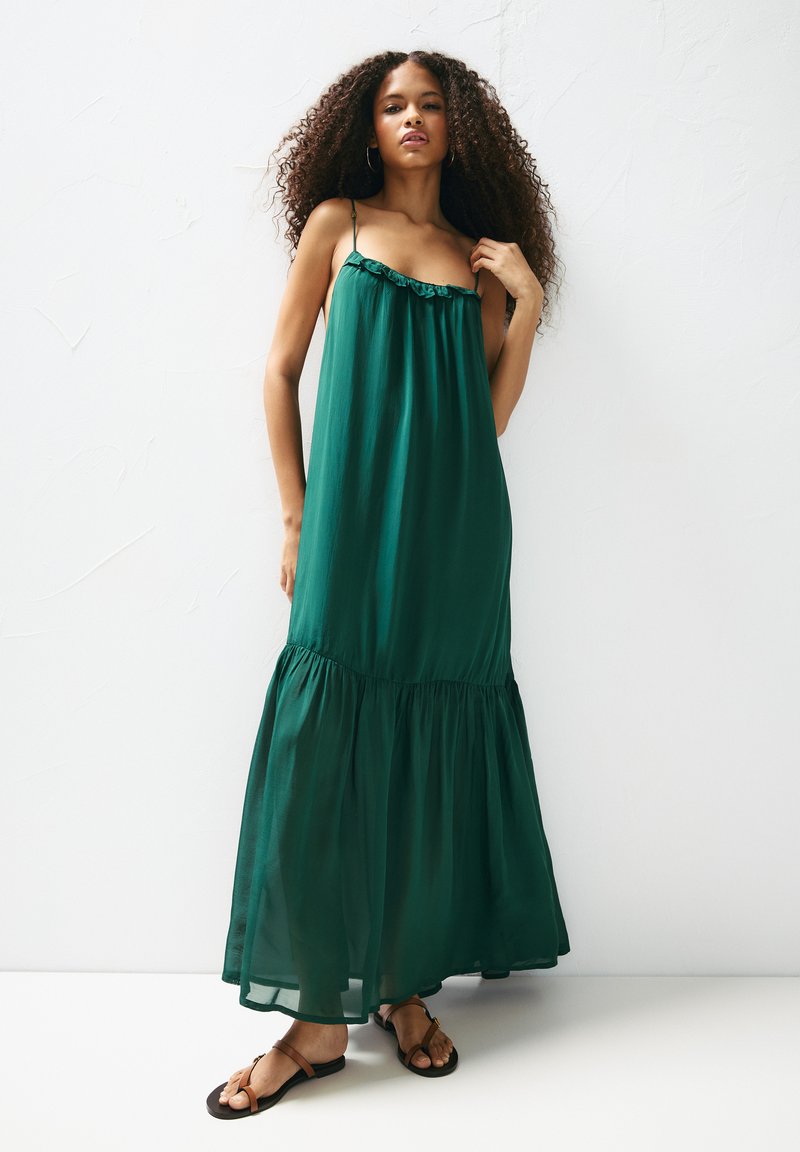 Rochie maxi verde, fluidă, cu bretele subțiri, decolteu volanat și fustă etajată. Stilizată cu sandale plate, așezată pe un fundal alb simplu.